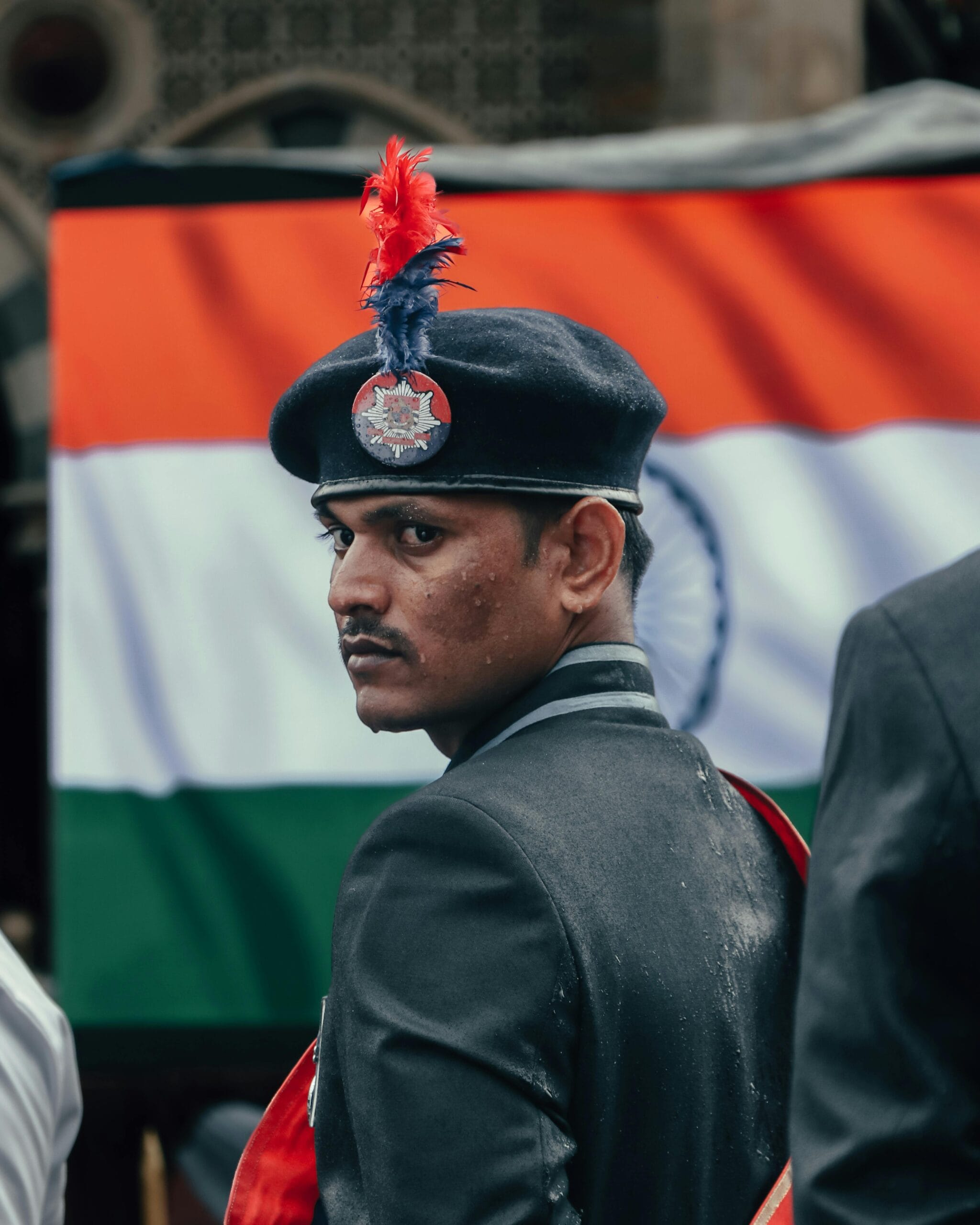 A solemn Indian soldier in ceremonial uniform with a flag background, outdoors.